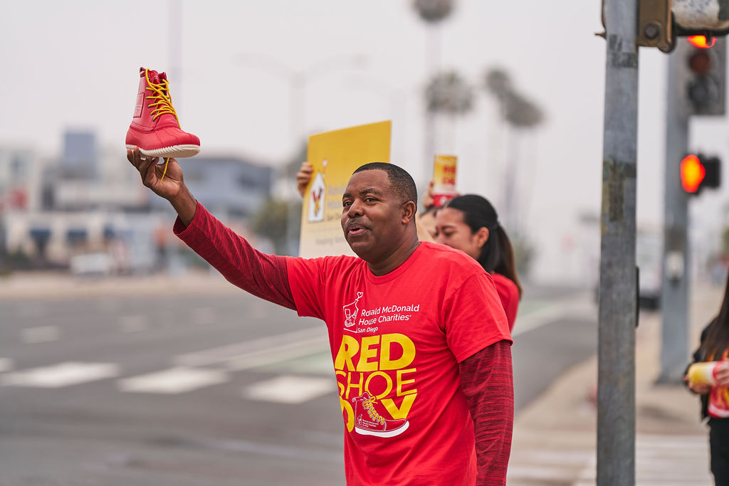 Red Shoe Day volunteer in a red shirt holds up an oversized red shoe to collect donations at a San Diego intersection.
